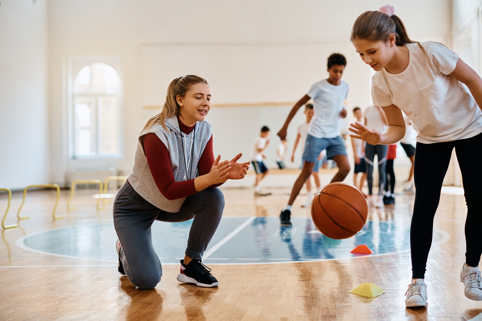 Schoolgirl leading a basketball among cones while sports teacher is supporting her during physical education class.